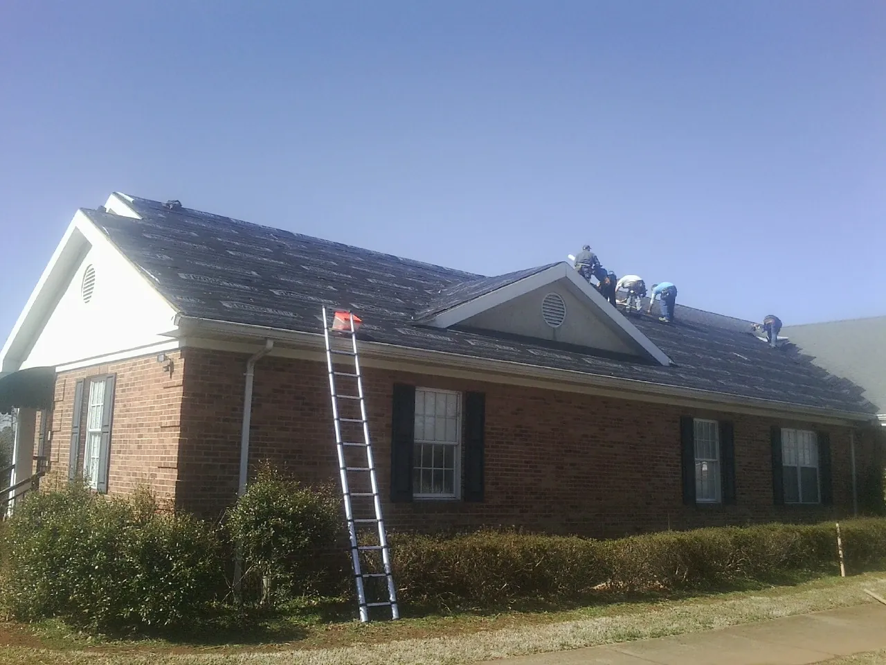Crew tearing shingles off a brick ranch roof with a ladder set — Metal Roof Repair work by our crew in Seth
