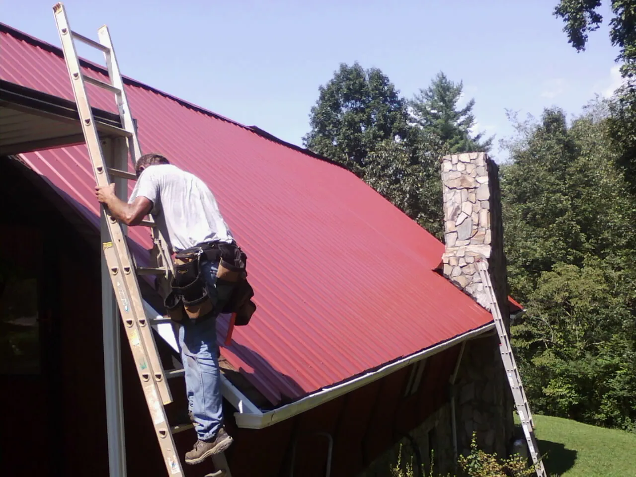 Roofer on a ladder beside a red metal roof and stone chimney — Soffit & Fascia Repair by Roofing Brothers in Seth